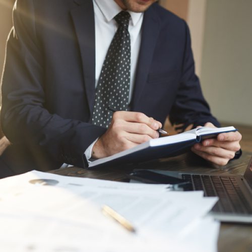 Closeup portrait of unrecognizable successful businessman wearing black formal suit  writing in planner while working with documentation and laptop